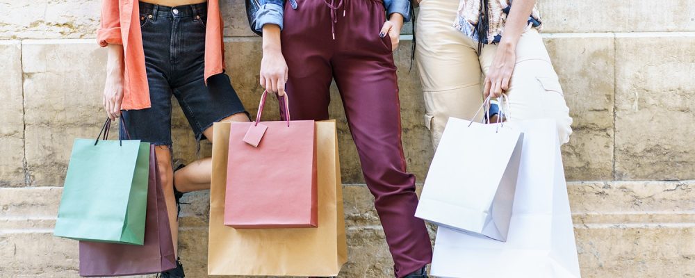 Crop anonymous stylish young female shopaholics in trendy outfits standing near aged stone building with shopping bags after successful purchases in city